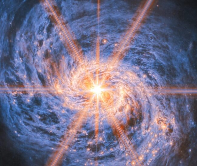 A spiral galaxy shown in mid-infrared light. The image is dominated by an extremely bright glow from the galaxy’s nucleus. Six large and two smaller rays of light emit from the centre, which are diffraction spikes created by the telescope’s optics. The galaxy’s spiral arms are visible by two lines of glowing orange bubbles which whirl out into the disc. Swirling blue clouds of dust make up the rest of the galaxy.